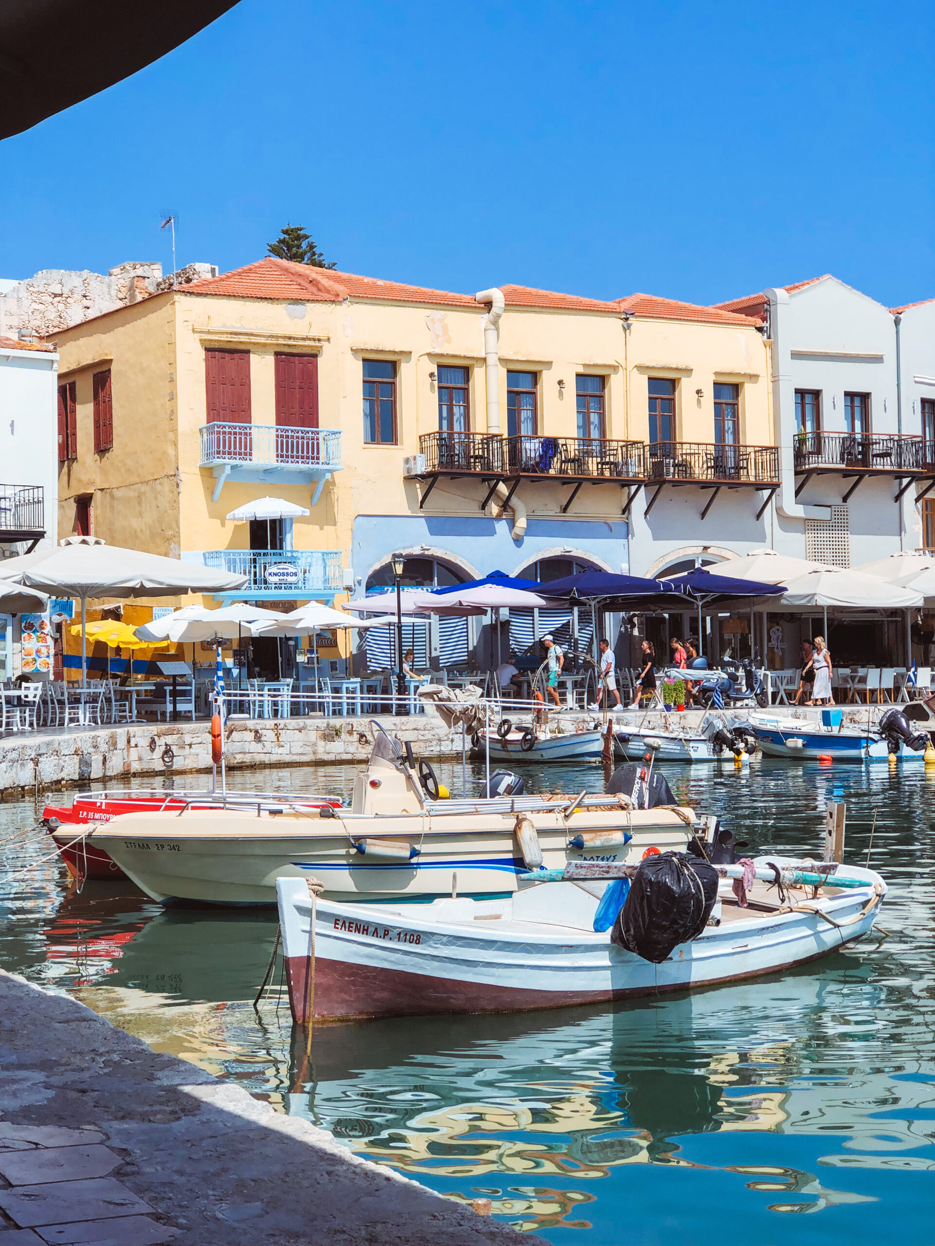 Scenic view of Rethymno’s Old Harbor in Crete with colorful fishing boats and historic waterfront buildings under a clear blue sky.